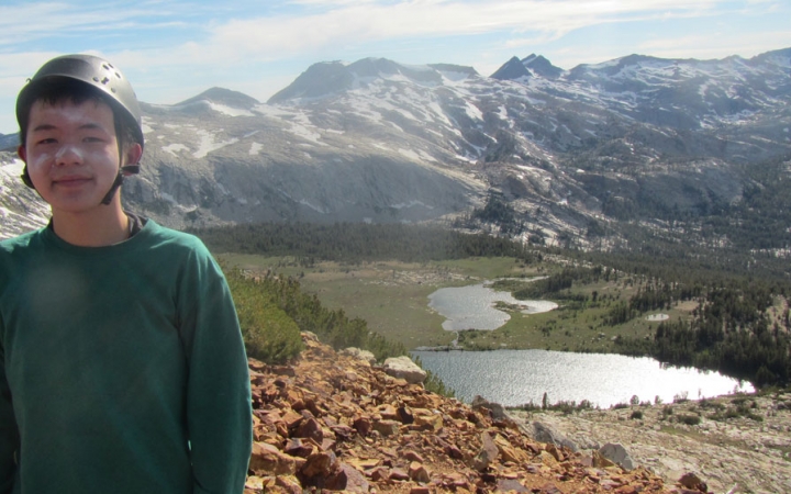 a person wearing a helmet and sunscreen poses for a photo in front a body of water and mountainous landscape